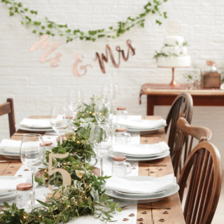 Houten tafel versierd met bladgroen, witte borden en wijnglazen en houten tafelnummers en hartjesconfetti