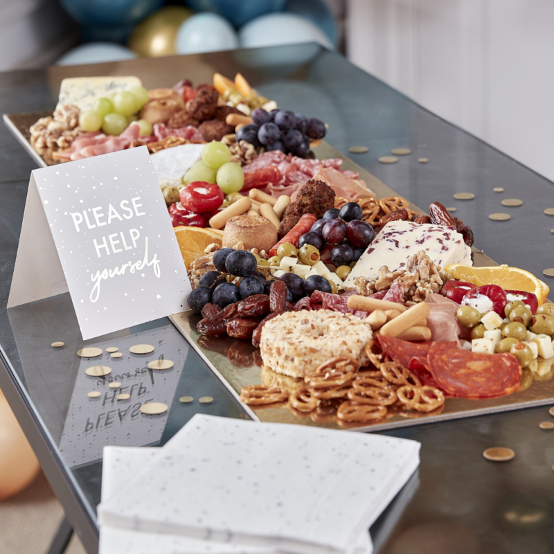 Gouden hapjesplank gevuld met olijven, pretzels, druiven en dadels ligt op een reflecterende tafel omringd door gouden confetti