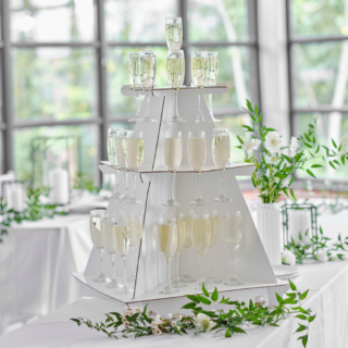 Witte drankjesstandaard met drie verdiepingen gevuld met champagneglazen staat op een witte tafel met witte bloemen en groene takken met bladeren