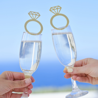 Champagneglazen worden vastgehouden voor een blauwe zee en zijn versierd met een gouden drankjestopper in de vorm van een ring