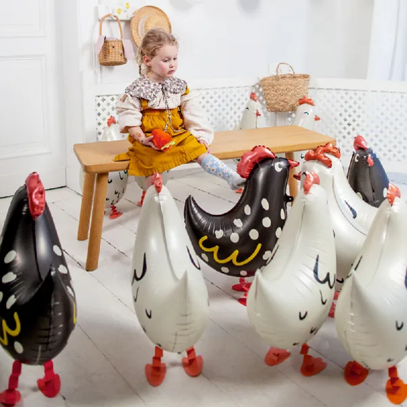 Meisje zit op een houten bankje in een kamer met folieballonnen in de vorm van kippen in het wit en zwart