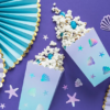 Popcornbakjes gevuld met popcorn liggen op een paarse tafel versierd met iridescent confetti en een lichtblauwe waaier met gouden rand