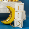 Witte blokken met gouden letters EID staan op een blauwe ondergrond naast een gouden ballon in de vorm van een halve maan