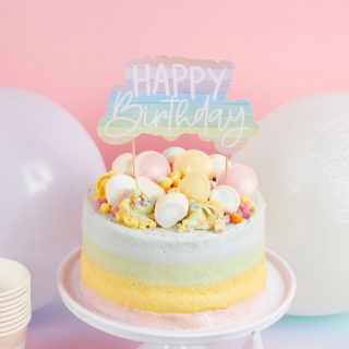 Taart topper met de tekst happy birthday in pasteltinten zit in een pastelkleurige regenboog taart op een wit plateau en staat voor ballonnen