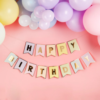 Pastelslinger met de gouden tekst happy birthday hangt voor een lichtroze muur onder ballonnen in pasteltinten