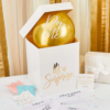 Witte doos met gouden tekst en metallic gouden ballon staat voor een roze muur op een witte tafel met blauwe en roze confetti