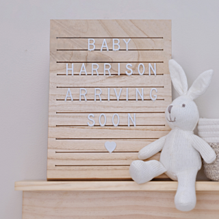 houten letterbord met witte letters staat op een plank in een babykamer naast een mandje en een wit knuffel konijn