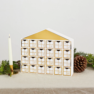 Advent kalender in het wit met goud in de vorm van een huisje staat op een jute kleed naast een kaars en een dennenappel