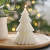 Kaars in de vorm van een witte kerstboom staat op een houten plateau voor een kerstboom met lampjes