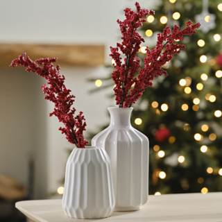 Witte vaasjes versierd met takken met rode besjes staan voor een verlichte kerstboom