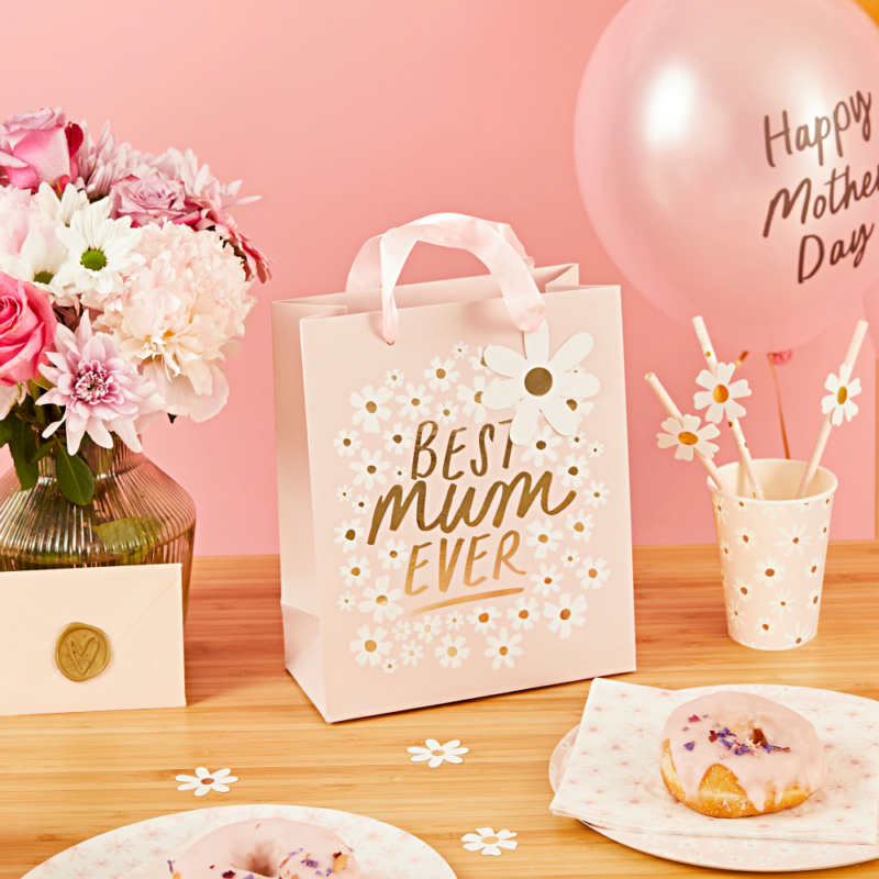Roze cadeautasje met de gouden tekst best mum ever en madeliefjes staat op een houten tafel naast een bosje met roze bloemen