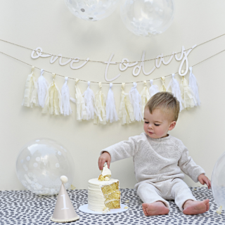 Jongetje zit op zijn eerste verjaardag bij een taart tussen de witte en creme versiering met confetti ballonnen en een tassel slinger