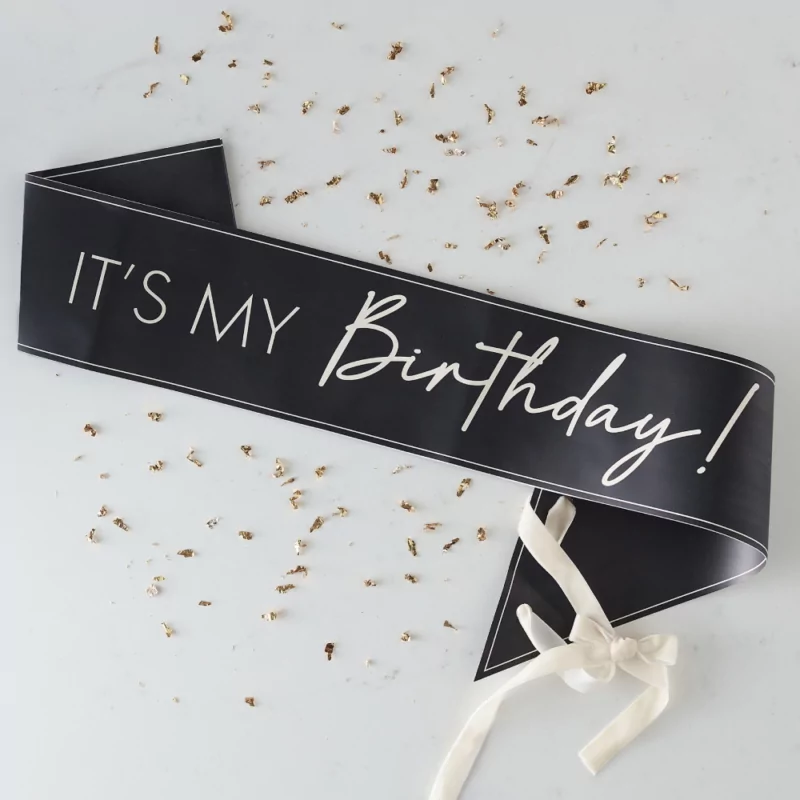 Zwarte sjerp met nudekleurige tekst it's my birthday! en een velvet lint ligt op een witte ondergrond omringd door gouden confettisnippers