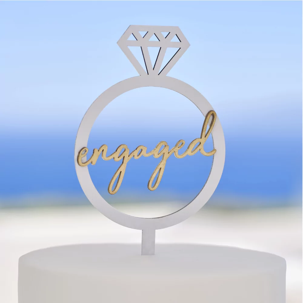 Witte taarttopper in de vorm van een ring met gouden woord engaged zit in een witte taart en staat voor een blauwe achtergrond