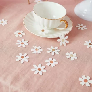 Tafelconfetti in de vorm van witte madeliefjes met een gouden binnenkant ligt op een roze linnen kleed naast een wit kopje met gouden handvat