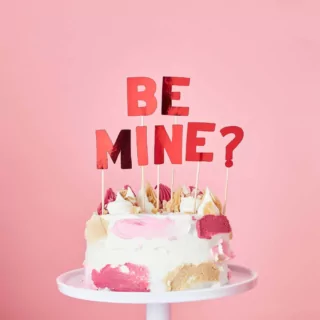 Rode taarttopper met de tekst be mine? zitten in een witte taart voor een lichtroze achtergrond