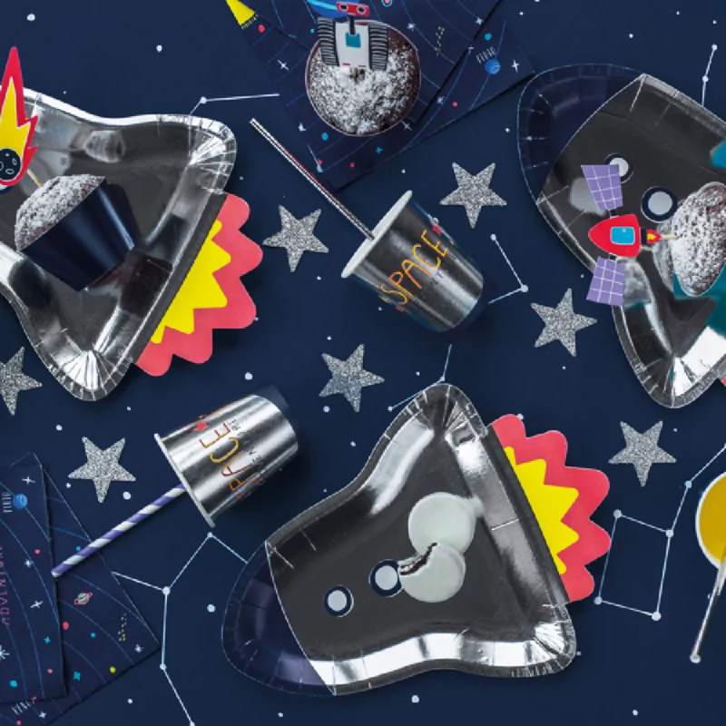 Donkerblauwe achtergrond met zilveren sterren is bedekt met metallic papieern bordjes in de vorm van een raket en metallic bekers met zilveren rietjes