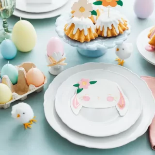 Lichtblauw tafelkleed bedekt met witte borden en servetten in de vorm van konijntjes, pastelkleurige kaarsjes in de vorm van paaseieren en witte kuikentjes