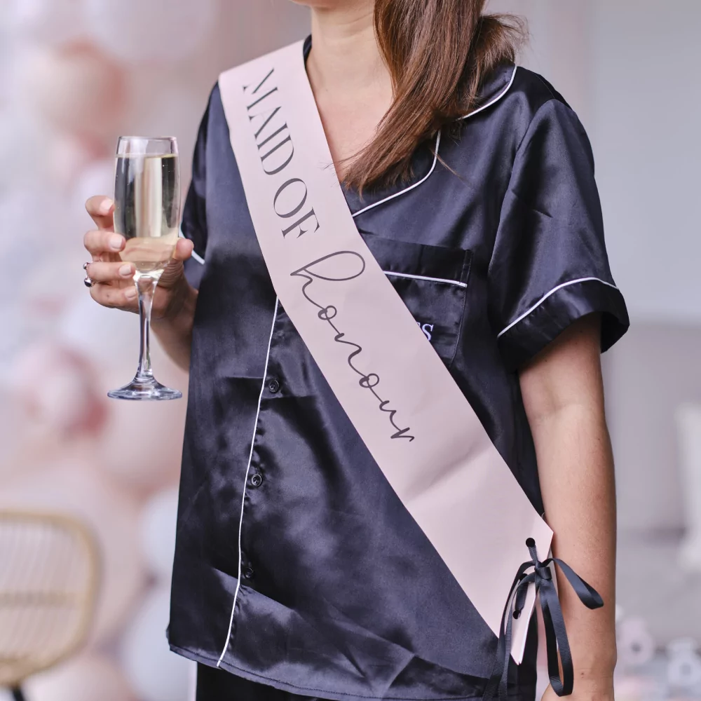 Vrouw in een donkerblauwe pyjama met een glas champagne draagt een roze sjerp met de zwarte tekst maid of honour