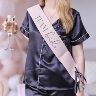 Vrouw in donkerblauwe pyjama draagt een roze sjerp met de tekst team bride en heeft een champagneglas vast