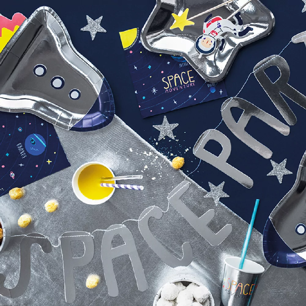 Zilveren slinger met de tekst 'spaceparty' ligt op een donkerblauwe ondergrond versierd met ruimte servetten en papieren bordjes in de vorm van een raket en zilveren sterren