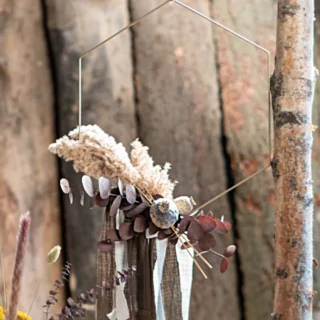 Gouden hanger in de vorm van een hexagon hangt boven een bosje droogbloemen naast een dunne boomstam