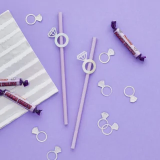 Paarse rietjes met diamanten ringetjes liggen op een lila ondergrond omringd door confetti met iridescent effect