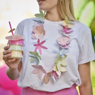Vrouw met wit shirt en roze broek heeft een bloemenkrans om en een beker met franjes in het roze, groen en geel