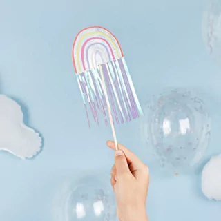 Toverstokje met regenboog en iridescent tassels wordt vastgehouden voor een lichtblauwe achtergrond naast confettiballonnen