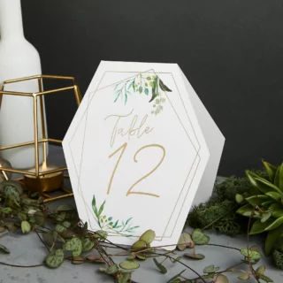 Tafelkaartjes met gouden tekst, groene bladeren en een geometrische vorm staan voor een zwarte muur naast een gouden kaarshouder