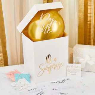 Witte doos met gouden tekst en metallic gouden ballon staat voor een roze muur op een witte tafel met blauwe en roze confetti