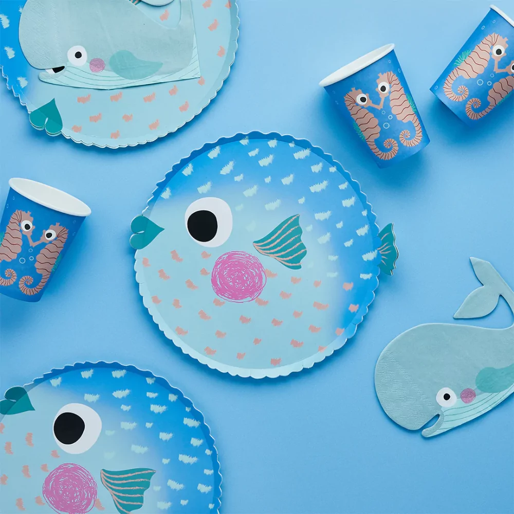 papieren bordje in de vorm van een kogelvis in het blauw met ombre effect ligt op een blauwe achtergrond naast walvis servetten en bekers met zeepaardjes