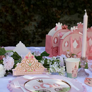Tafel versierd met een roze tiara, bordjes met een koets erop en cadeautasjes in de vorm van een koets naast een bloemenslinger en een lange kaars