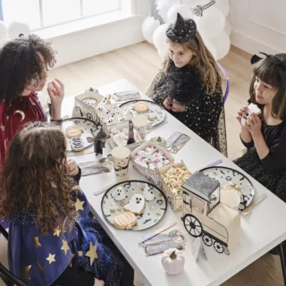 Kinderen in halloween kostuums zitten aan een tafel versierd met een treintje met snacks, bordjes en bekers met halloween poppetjes erop