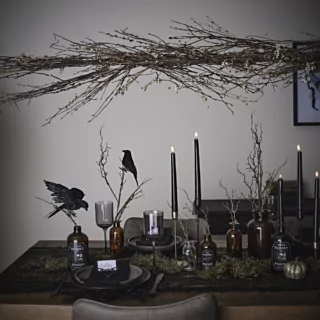 Pronkstuk met bruine takken en groene en witte bessen hangt boven een donkerbruine tafel versierd met kraaien en zwarte kaarsen voor een grijze muur