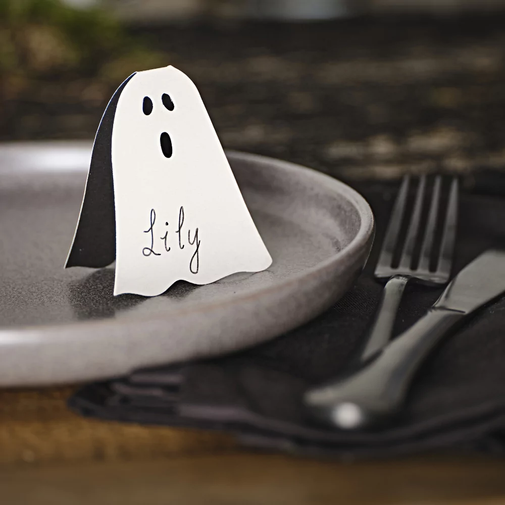 Tafelkaartjes in de vorm van een spookje met gezichtje staat op een zwart bord, naast zwart bestek op een zwarte, stoffen servet