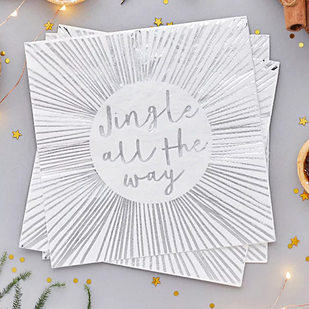 Witte servetten met zilveren strepen en de tekst jingle all the way liggen naast gouden confetti en groene takjes van een dennenboom