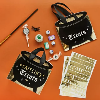 Cadeautasjes in de vorm van een zwart keteltje met gouden stickers om het te personaliseren liggen op een oranje ondergrond