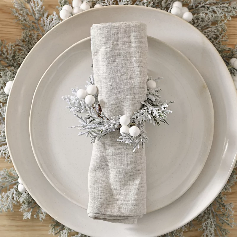 Servettenring met groene, besneeuwde takjes met witte besjes zit om een beige, stoffen servet heen en ligt op een wit bord op een houten tafel