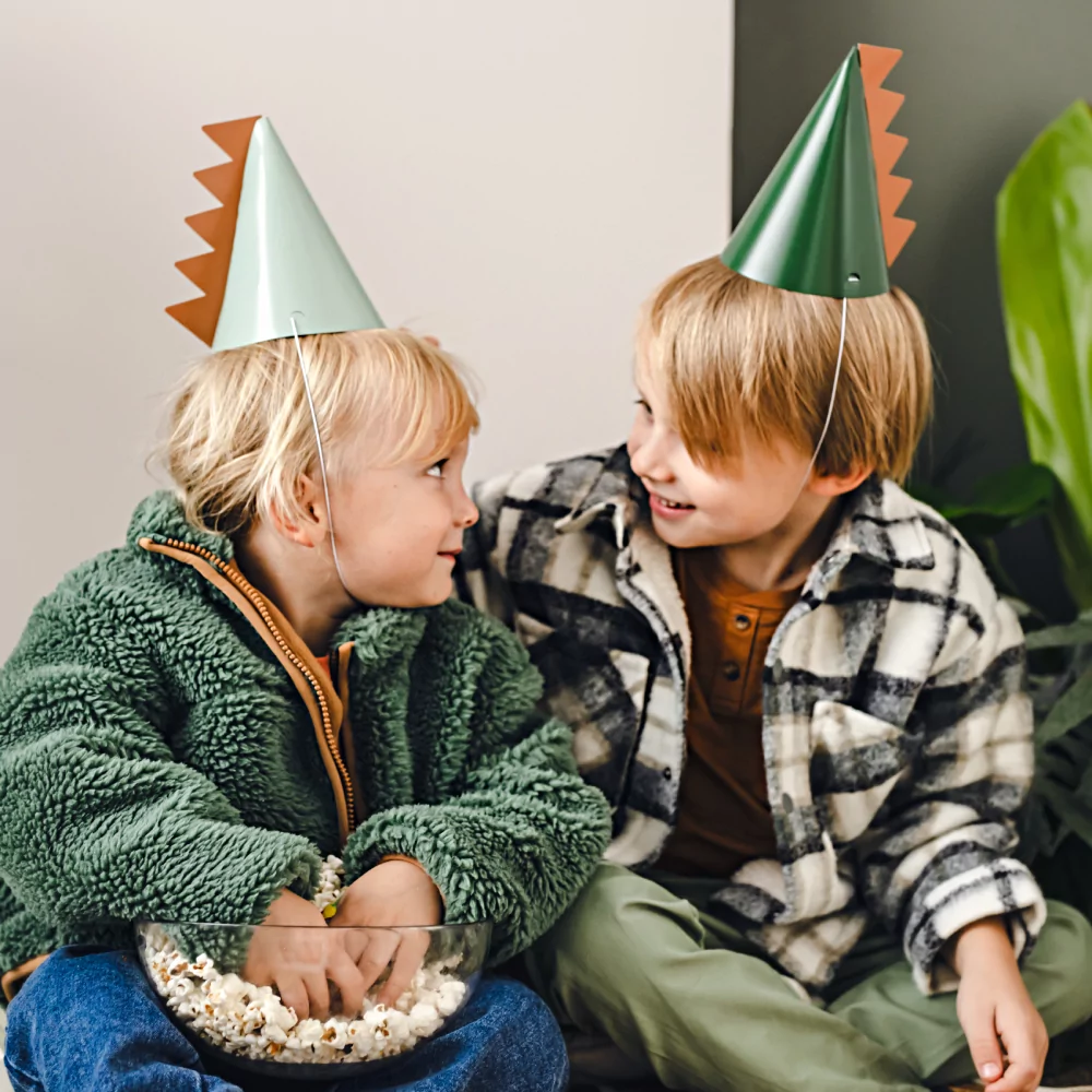 Feesthoedjes in het bruin en groen met een dino staart worden gedragen door twee jongetjes op een dino kinderfeestje