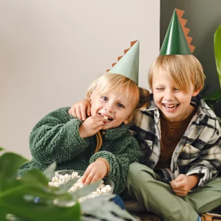 Feesthoedjes in het bruin en groen met een dino staart worden gedragen door twee jongetjes op een dino kinderfeestje