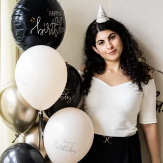 vrouw met een feesthoedje op en een bundel zwarte en creme ballonnen met de tekst happy birthday