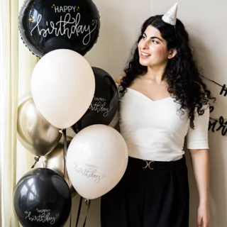 vrouw met een feesthoedje op en een bundel zwarte en creme ballonnen met de tekst happy birthday