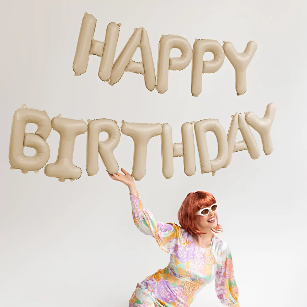 beige ballonnen met de tekst happy birthday hangen boven een vrouw met rood haar