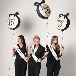 drie vrouwen in zwarte pakken met ballonnen in het creme en zwart met de teksten happy 18, happy 40 en happy 60